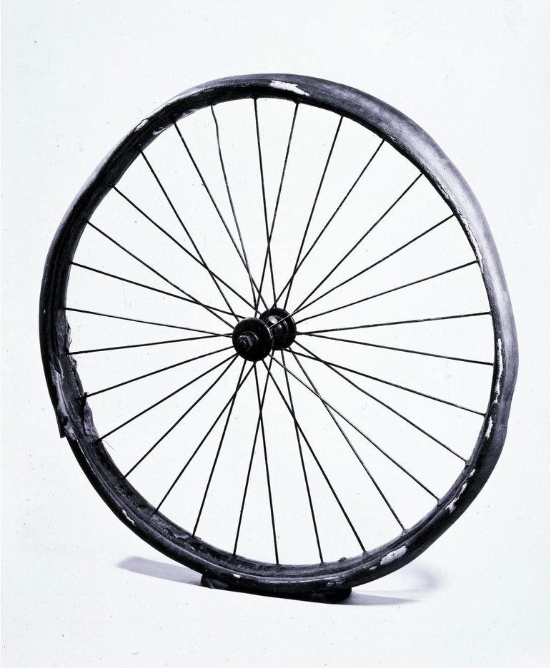 A colour photograph of a large bicycle wheel with 30 spokes. It is quite weathered. The rim is surrounded by a degraded rubber.
