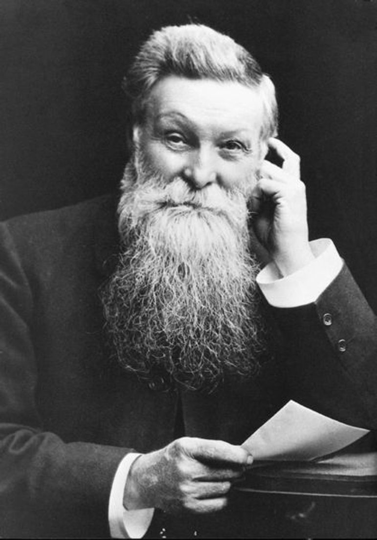 A black and white photograph of a man in a classic Victorian portrait pose. He has thick swept-back hair with a left side parting, and a very long beard. He's wearing a Victorian double-breasted coat. His left hand is touching the left side of his face and his left elbow is resting on a surface. His right hand is holding a piece of paper at mid level.