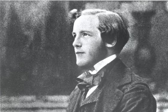 A black and white photograph of a young man wearing a white collared shirt, dark suit jacket and waistcoat, sitting on a bench holding a spinning top.