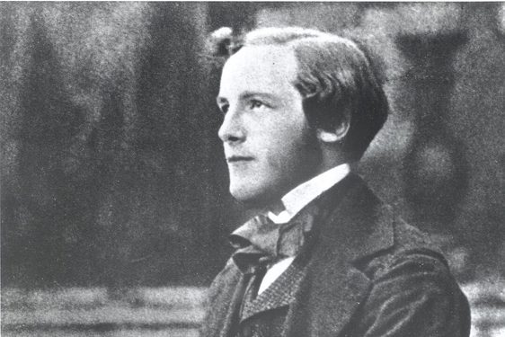 A black and white photograph of a young man wearing a white collared shirt, dark suit jacket and waistcoat, sitting on a bench holding a spinning top.