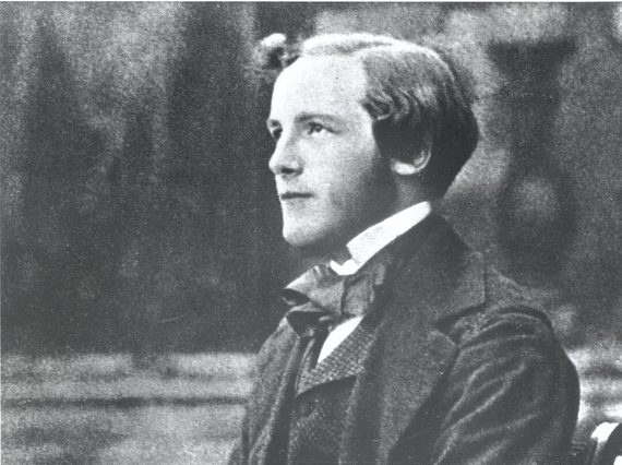 A black and white photograph of a young man wearing a white collared shirt, dark suit jacket and waistcoat, sitting on a bench holding a spinning top.