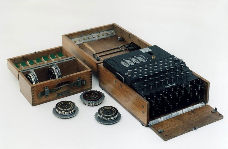 A colour photograph of a 4-rotor enigma machine in a wooden box and another smaller wooden box with 5 extra rotor wheels. The machine is black and has a number of keys on the top front and many jack sockets at the front edge with number and wires attached to some