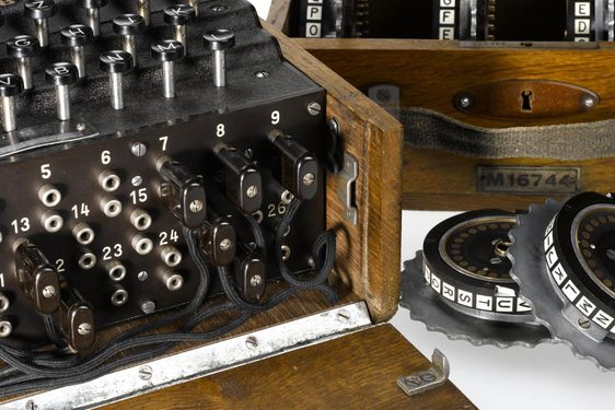 A colour photograph of a close-up of the 4 rotor enigma machine showing the front which has Manu jack sockets on the front side with numbers and wires attached to some and a keyboard on the top. There is a separate box with extra rotor dials which are metal and circular.