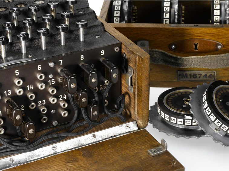 A colour photograph of a close-up of the 4 rotor enigma machine showing the front which has Manu jack sockets on the front side with numbers and wires attached to some and a keyboard on the top. There is a separate box with extra rotor dials which are metal and circular.