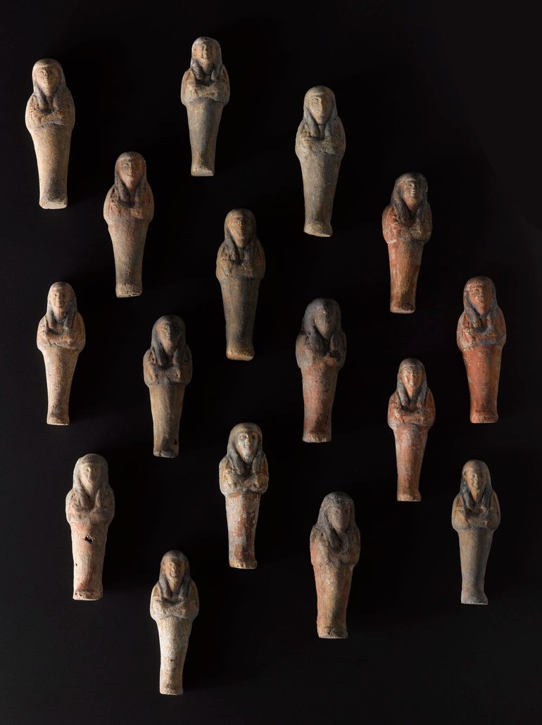 Sixteen plain pottery mould-made figures in the shape of a mummified person, spread out against a black background.