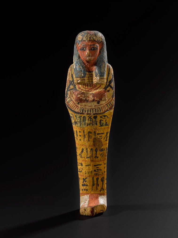 A brightly painted wood figure in the shape of a mummified person, with five lines of hieroglyphics written on it. The figure is against a black background.