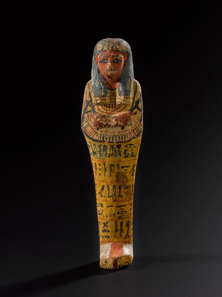 A brightly painted wood figure in the shape of a mummified person, with five lines of hieroglyphics written on it. The figure is against a black background.
