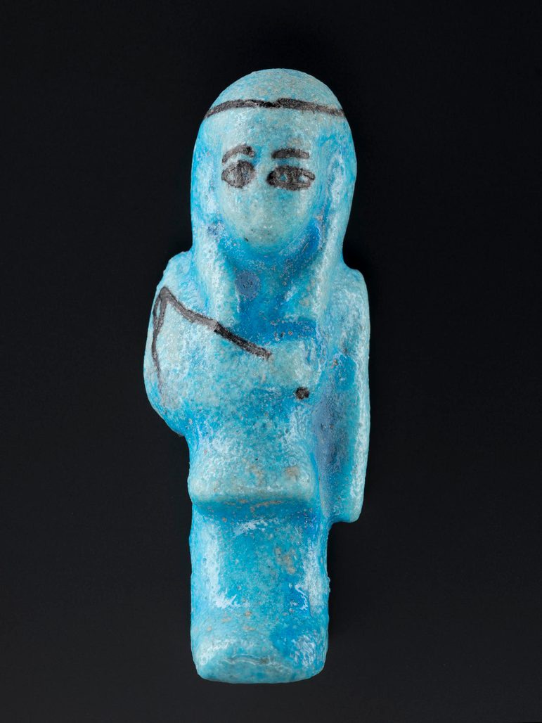 A ceramic blue figure made in the shape of a mummified person with outlined facial features and a whip in black. The figure is against a black background.
