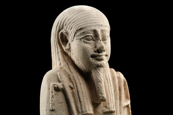 A cream-coloured mould-made ceramic figures in the shape of a mummified person, surrounded by a black background.