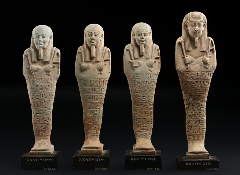 Four cream-coloured mould-made ceramic figures in the shape of a mummified person, spread out in a line against a black background.