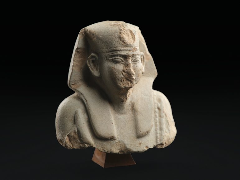 The head and shoulders of a faded cream coloured ceramic figure in the shape of a mummified person, wearing an Ancient Egyptian headdress. The figure is against a black background.bol and a sceptre. The figure is against a black background.
