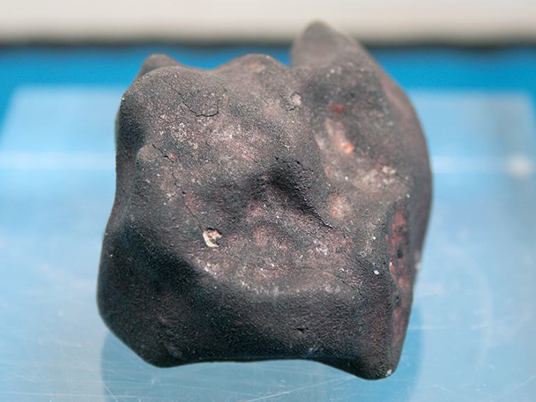 A grey meteorite with pinkish brown flecks sitting on a blue surface