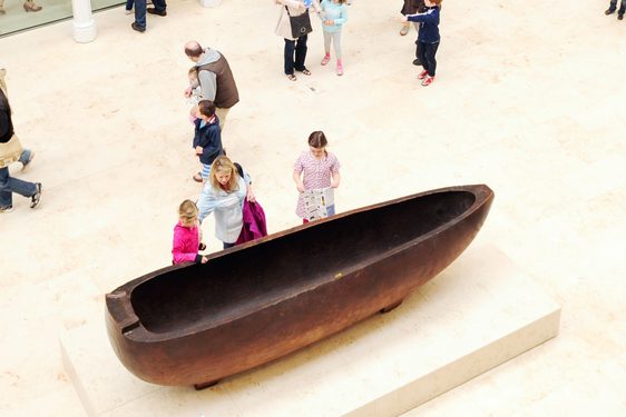 A large oval shaped wooden object with a hollowed out middle on display in a museum with people looking at it