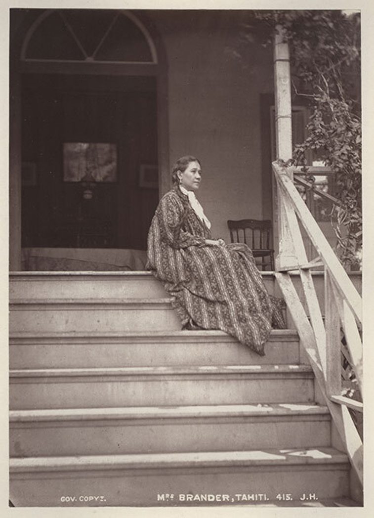 A black and white image of a woman in a long patterned dress wearing her hair in a long dark braid, sitting on stairs that lead to a house entryway.