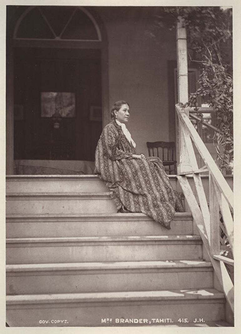 A black and white image of a woman in a long patterned dress wearing her hair in a long dark braid, sitting on stairs that lead to a house entryway.