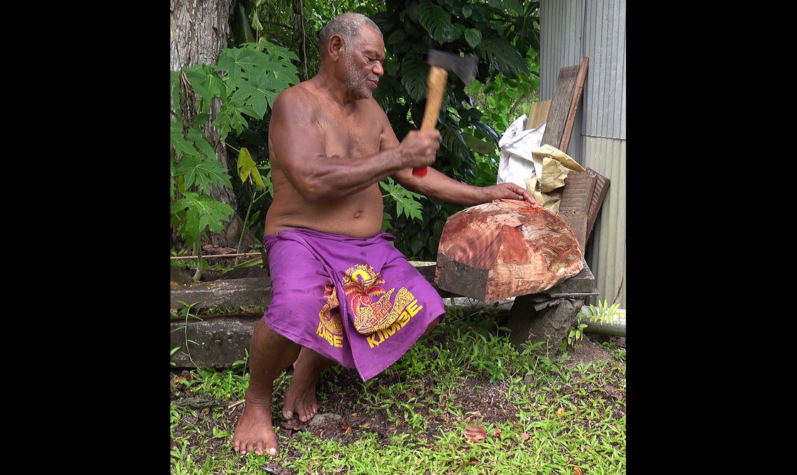 A man sitting on a bench prepares to hit a block of wood with an axe.