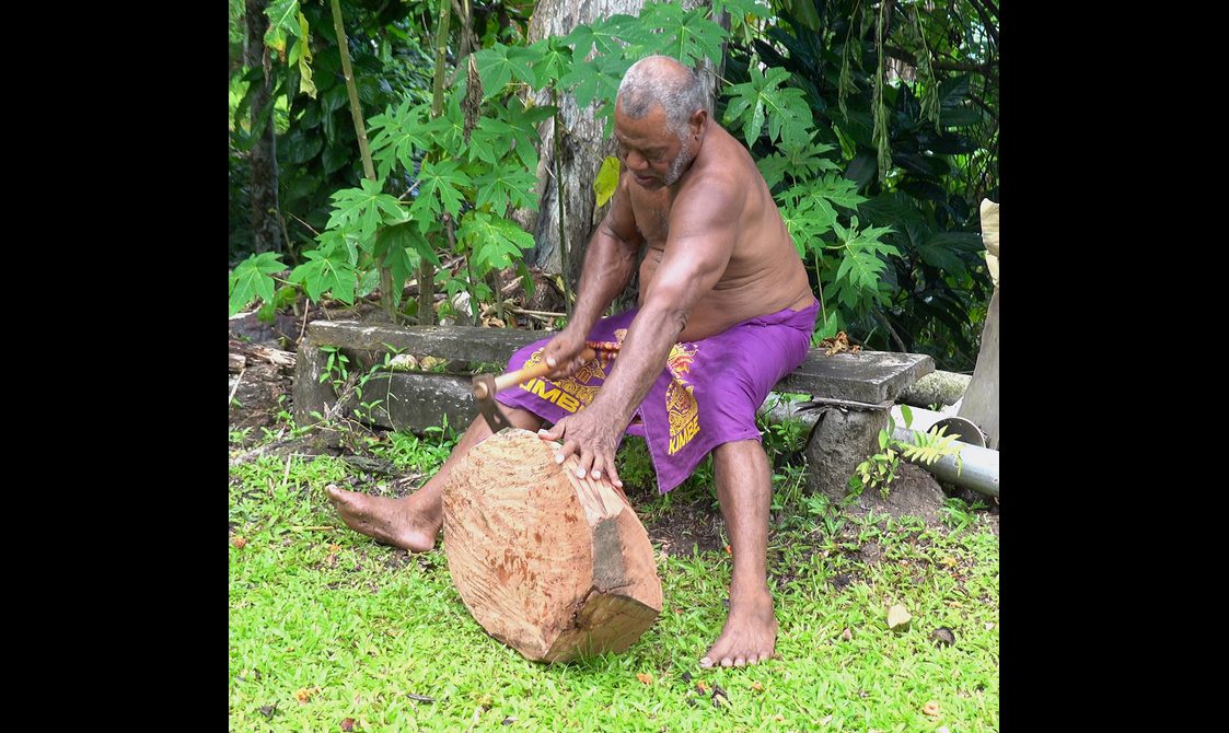 A man sat on a bench beginning to work a slab of wood with a tool.