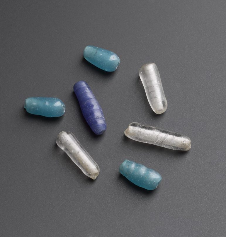 A selection of long tapered glass beads on a grey background. The beads are turquoise, blue, or opalescent in colour and arranged randomly.