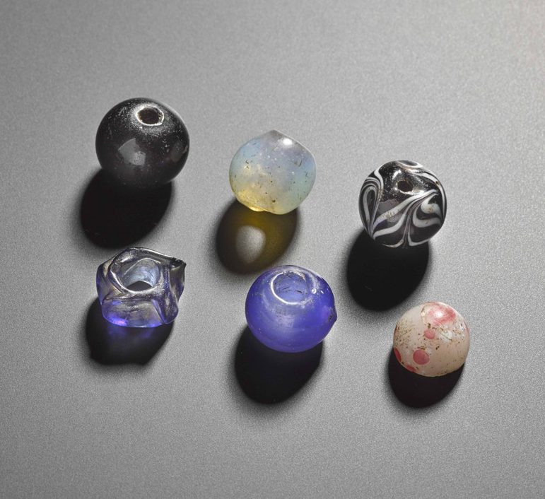 A collection of 6 beads of varying colours against a great background. There is a ball brand, a clear opalescent bead, a mblack and white marble bead, a blueish opalescent doughnut-shaped jagged bead, a blue opalescent bead with a large central hole, and a smaller cream and brown marbled bead.