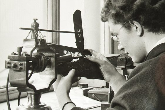 A black and white photograph of a woman to the right, with short wavy brown hair, wearing a lose back jacket at a studio desk. She is operating am old-fashioned lathe. There is a bright double window to left.