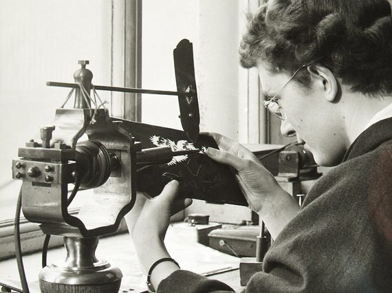 A black and white photograph of a woman to the right, with short wavy brown hair, wearing a lose back jacket at a studio desk. She is operating am old-fashioned lathe. There is a bright double window to left.