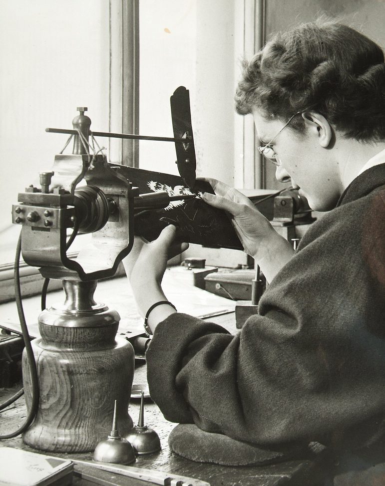 A black and white photograph of a woman to the right, with short wavy brown hair, wearing a lose back jacket at a studio desk. She is operating am old-fashioned lathe. There is a bright double window to left.