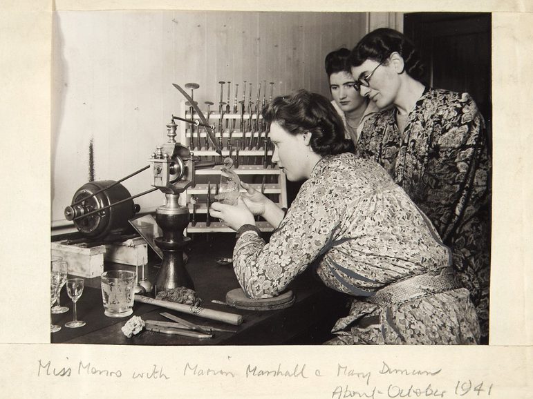 A black and white photograph of 3 women in 1940s flowery dresses and 40s hair styles in a studio. There is one woman seated and operating an old lathe. The other tow women are standing behind her watching as she is working.