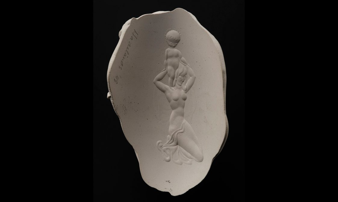 An oval plaster cast against a black background. There is an image of a stylised semi-naked woman holding a baby above her head.