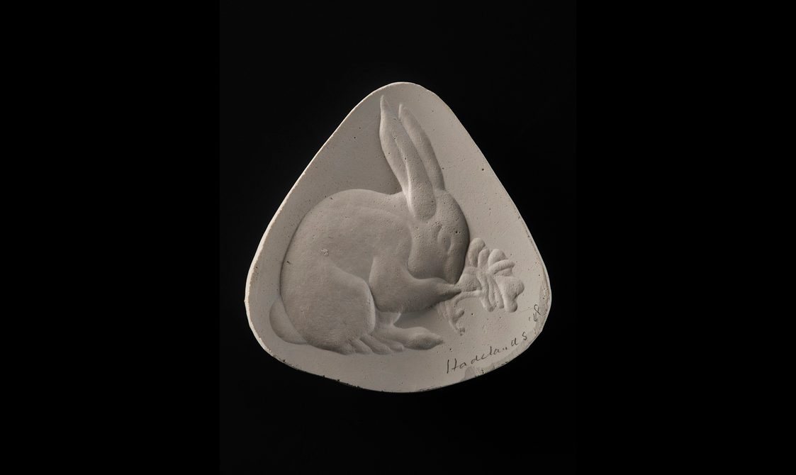 A triangular shaped plaster cast against a black background. There is a bas relief of a rabbit eating some vegetation, taking up most of the interior surface.