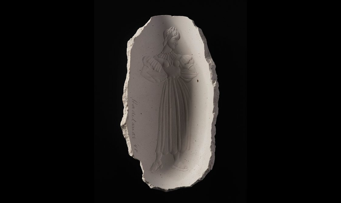 An oval plaster cast against a black background. The cast has begged edges and a bas relief of a stylised woman wearing a long classical gown.