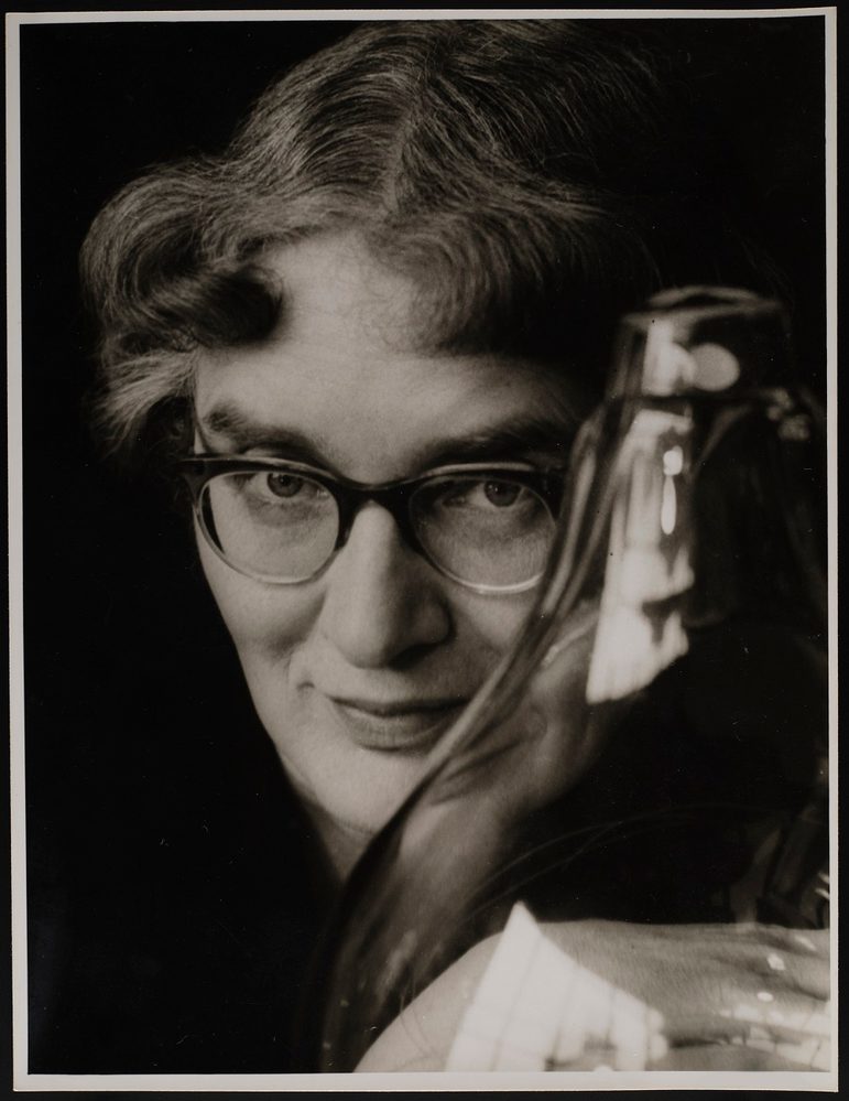 Close up black and white portrait of a woman with wavy grey hair and thick 50s style glasses against a black background. In front of her to the right is a large glass object. Behind it, her clasped hands can be seen,