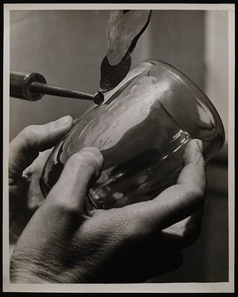 A cblack and white close up of two hands holding a drinking glass, The glass is held underneath a lathe machine.