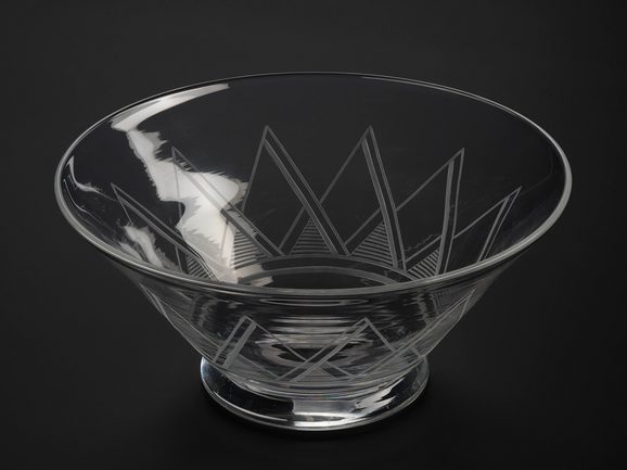 A tall wide glass bowl against a black background. The bowl is tall with a wide brim which tapers into a narrower base. The sides have a bold geometric triangular pattern around the bottom of the outside.
