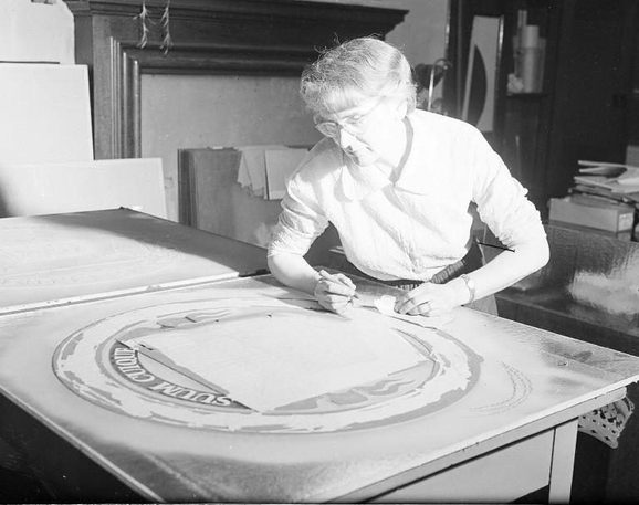 A black and white photograph of a woman with wshort white hair, glasses and a white short, leaning over a large circular glass sheet on a large table. Behind her are many frames and materials. The glass on the table has a circular design with lettering and patterns around the edge.