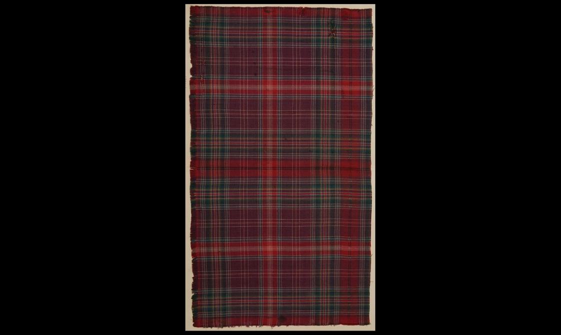 Tall rectangular swatch of burgundy, dark blue, red and light green tartan fabric against a black background.