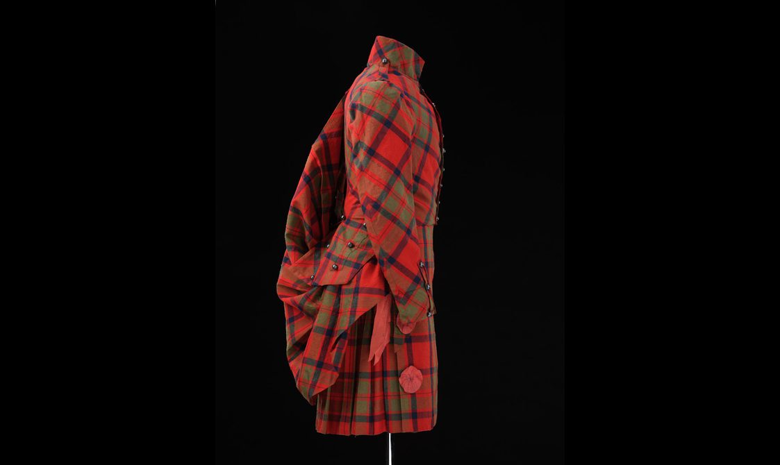 A side view of a mannequin wearing a red tartan kilt, jacket ,and shawl.