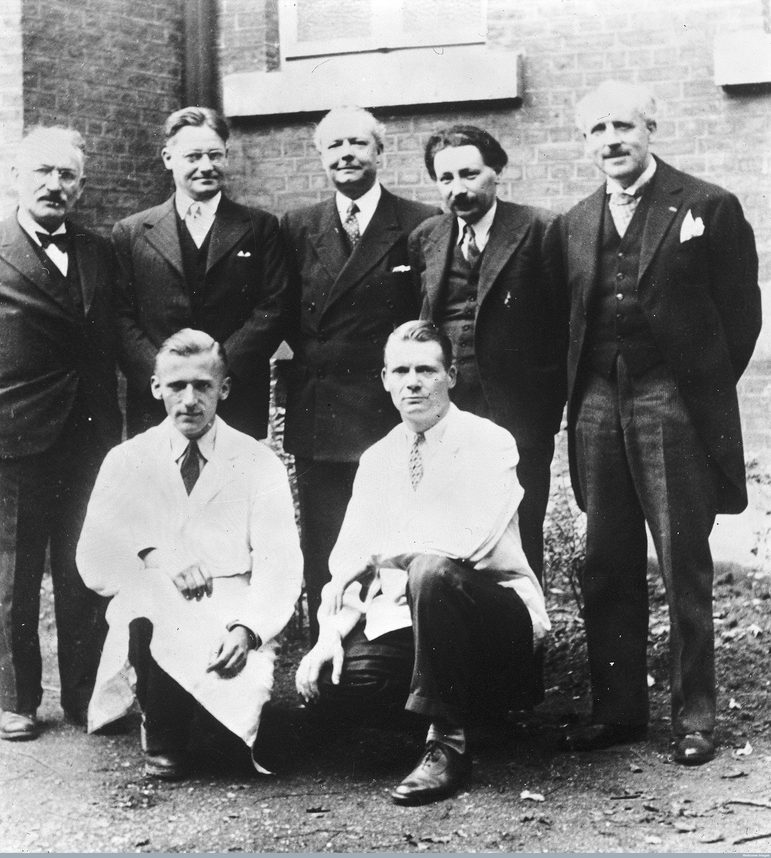 A black and white photograph of five men in dark suits standing behind two men in white lab coats.
