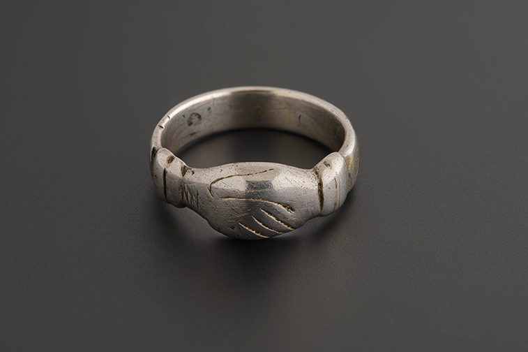 A small silver ring with clasped hands decoration and faint markings on the inside of the band, against a grey background.