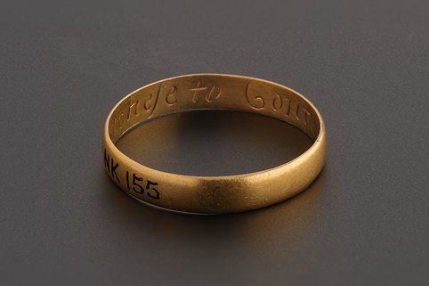 A gold posy ring with plain band. The word 'ese to content' of the phrase 'No Richese to content' can be seen inside the band.