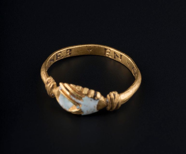 A gold ring with clasped hands, with flecks of white enamel. The ring is against a black background. The ring has an inscription: 'Prenes En Gre'.