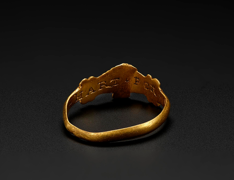 The back of a shiny gold ring, showing an inscription '+HART+FOR+♥+'. The ring is set against a black background.