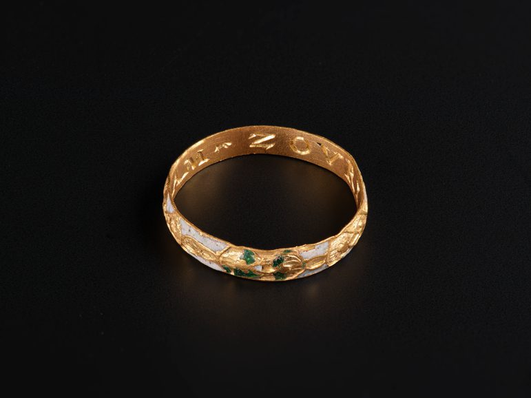 A small gold ring with a band decorated with green and white enamel against a black background. Inside there's an inscription: "+I+AM-ZOVRIS+".