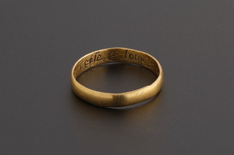 A simple gold ring against a dark grey background. A section of writing from the phrase 'lett murning cease and love increase' on the inside of the band.