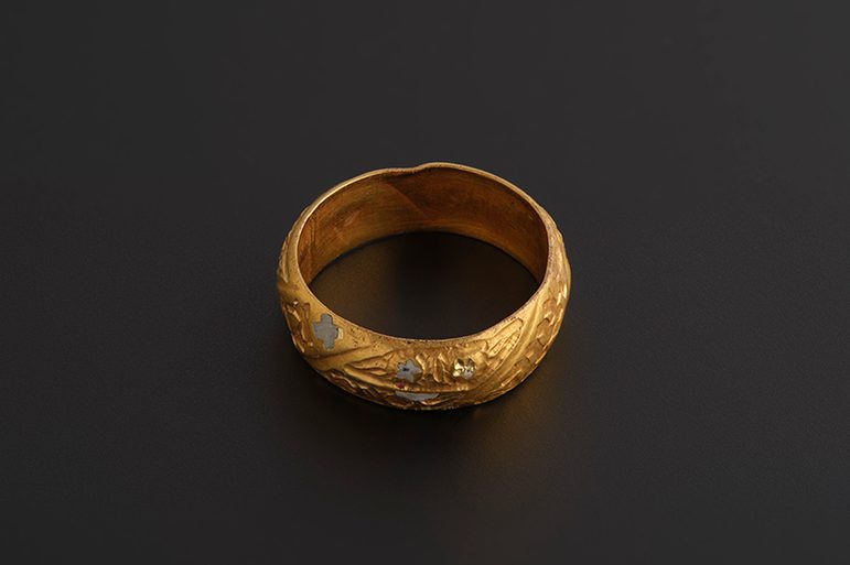 A textured gold ring with enamel flowers and markings against a dark grey background, with faint writing marks on the inside of the band..