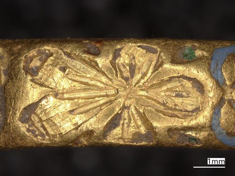 Microscopic close-up of small gold ring, showing decorative designs, small remains of white and green enamel and a floral design.