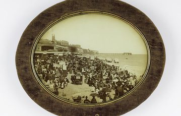 A black and white image of a group of people in Victorian dress with large cameras on a beach. The image is presented in a wooden oval frame.