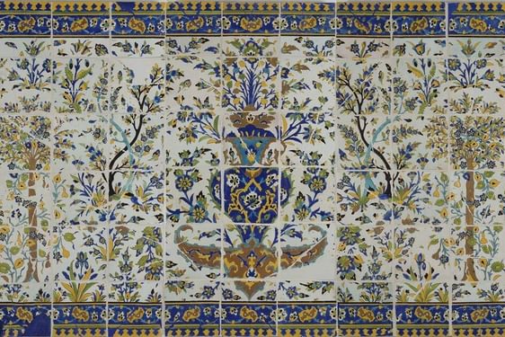 A long, rectangular panel made of hundreds of intricately decorated, painted tiles. This image gives the overall impression of the illustration, which from a distance is of a garden scene with trees all around, and a fountain as the centrepiece. The colours painted are yellow, blue, green and brown.