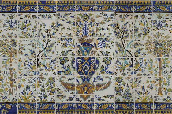 A long, rectangular panel made of hundreds of intricately decorated, painted tiles. This image gives the overall impression of the illustration, which from a distance is of a garden scene with trees all around, and a fountain as the centrepiece. The colours painted are yellow, blue, green and brown.