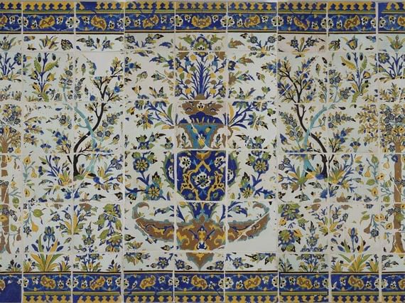 A long, rectangular panel made of hundreds of intricately decorated, painted tiles. This image gives the overall impression of the illustration, which from a distance is of a garden scene with trees all around, and a fountain as the centrepiece. The colours painted are yellow, blue, green and brown.