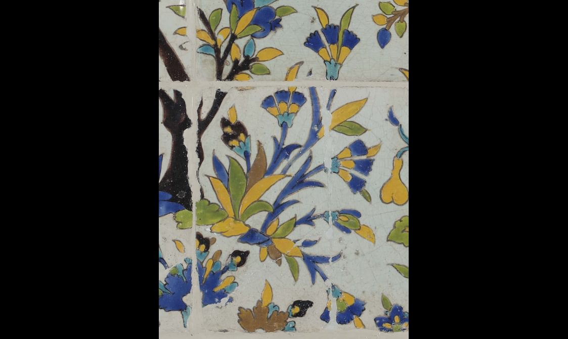 A section of a long, rectangular panel made of hundreds of intricately decorated, painted tiles. This section focuses on a small plant with flowers. The colours painted are yellow, blue, green and brown.